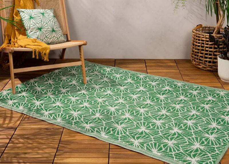 A polyester outdoor rug with a tropical leaf design, laid on a wood deck surface below an outdoor chair.