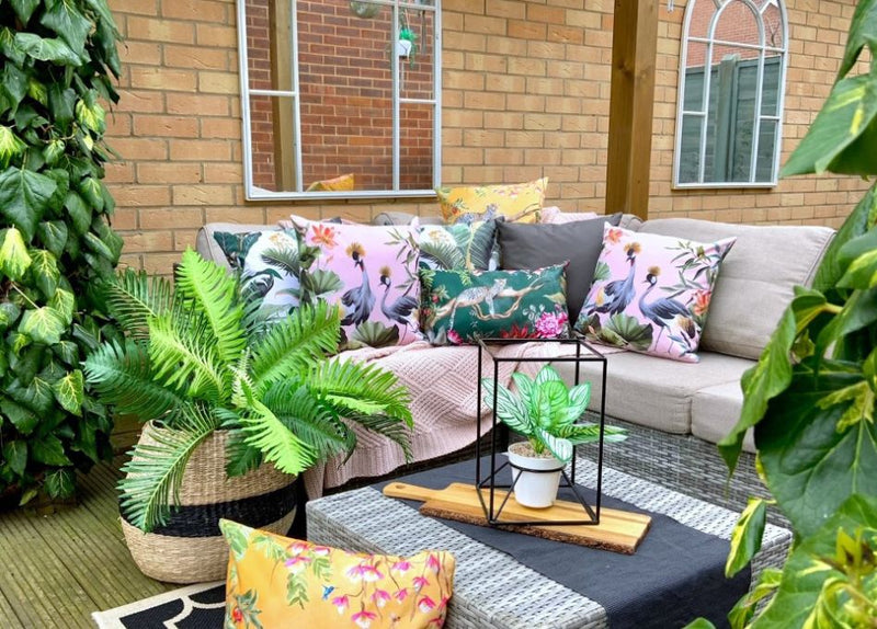 Four pink outdoor cushions of different sizes scattered on a floor and woven garden furniture, featuring summery patterns of citrus fruits.