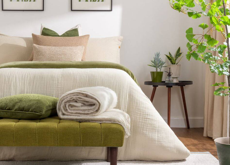 A biophilic bedroom design with neutral cotton muslin bedding, earthy beige and green scatter cushions, a moss bedspread, a white mohair throw and indoor plants.