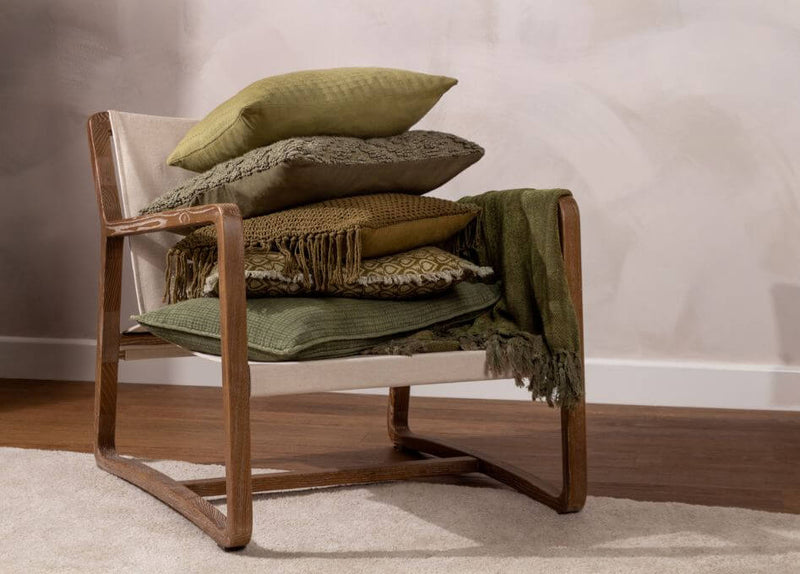 A selection of moss, olive and forest green cushions stacked on a wooden chair with a green throw draped over the arm, in a room with beige walls and a beige rug.