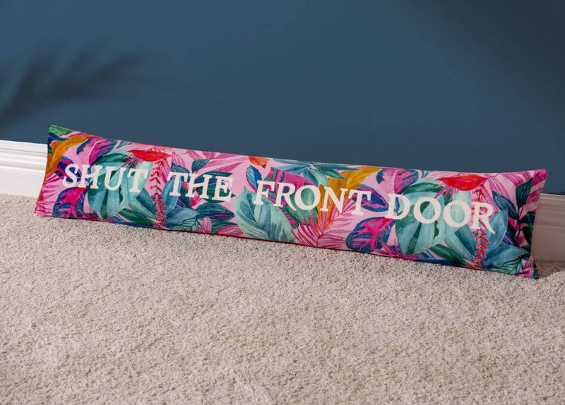 A vibrant draught excluder cushion with a multicoloured exotic leaf design and a printed slogan reading 'shut the front door', laid on a grey carpet in front of a dark blue wall.
