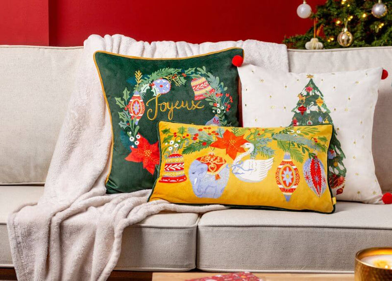 Three green, gold and white Christmas cushions with various festive designs, arranged on a neutral couch with a beige throw.
