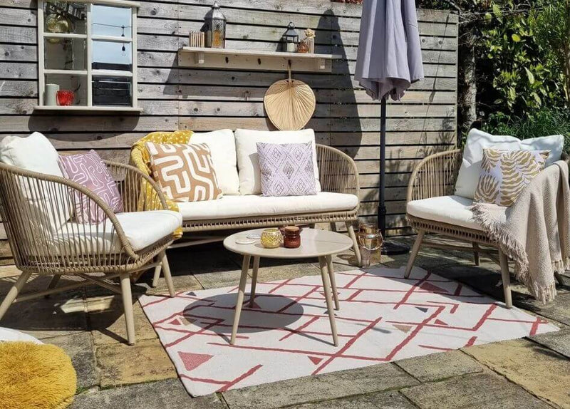 A garden scene of an outdoor rug placed on grass underneath a garden chair, an outdoor cushion and various summer ornaments.