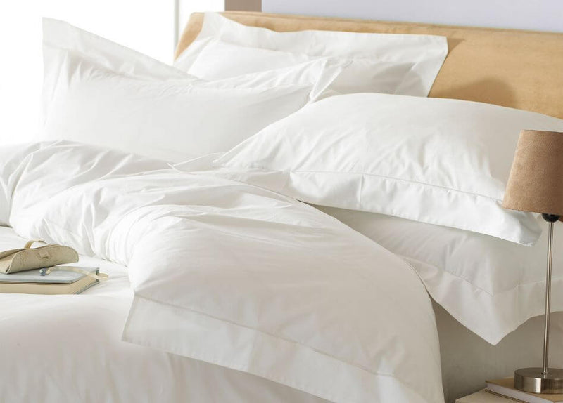 A white, 200 thread count, Oxford bordered 100% cotton duvet set with patching pillowcases, tousled on a bed with a beige fabric bedframe, with a woven side table holding books and a desk lamp.