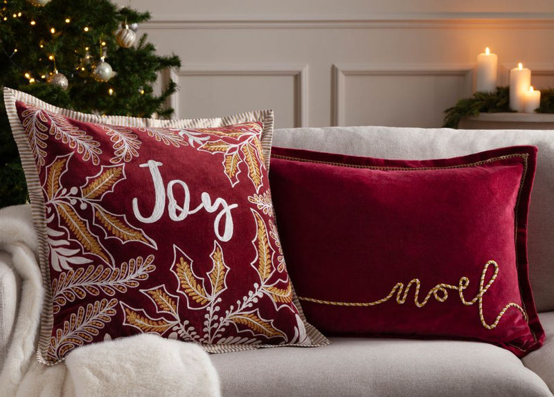 A sofa infront of a Christmas tree, with two Christmas cushion on it saying Joy and Noel