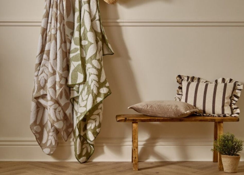 Two leaf woven throws in beige and olive green next to a wooden bench and a small potted plant with a plain and striped cushion on top