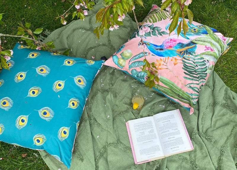five outdoor cushions with bright statement slogan designs, scattered on a grass surface.