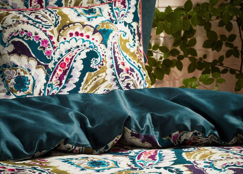 A teal paisley pillowcase on a folded over duvet cover set, in the background you can see a white painted brick wall that's got a green plant growing over it.