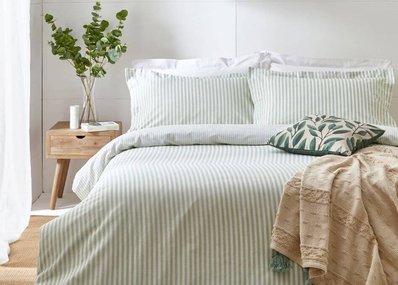 A white, 200 thread count, Oxford bordered 100% cotton duvet set with patching pillowcases, tousled on a bed with a beige fabric bedframe, with a woven side table holding books and a desk lamp.