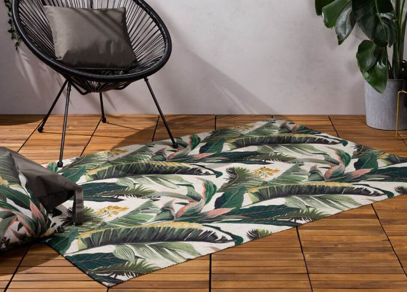 A polyester outdoor rug with a tropical leaf design, laid on a wood deck surface below an outdoor chair.
