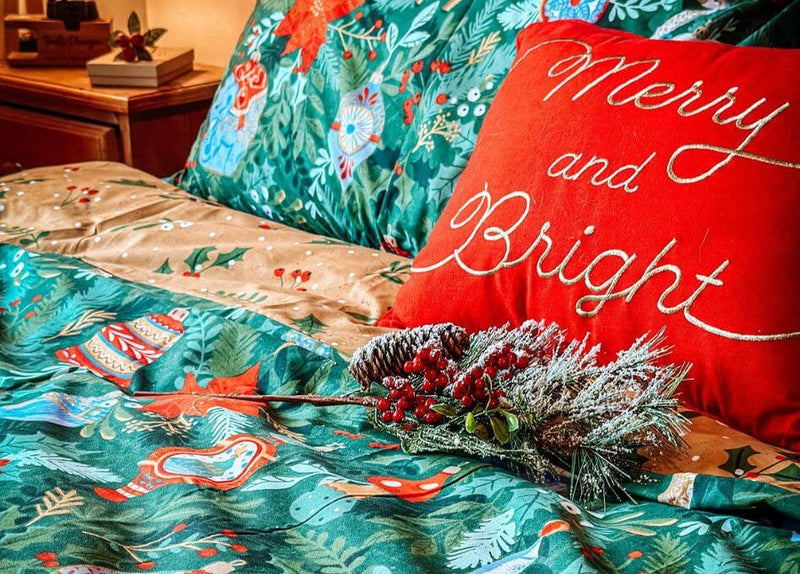 A green festive duvet cover set with a traditional design of baubles and trinkets, arranged on a bed with a red scatter cushion and some festive foliage.