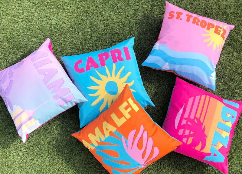 five outdoor cushions with bright statement slogan designs, scattered on a grass surface.
