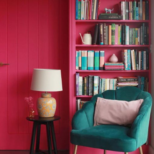 A cosy reading nook with hot pink walls, a hot pink bookshelf, an emerald green chair and a yellow table lamp stood on a small side table.