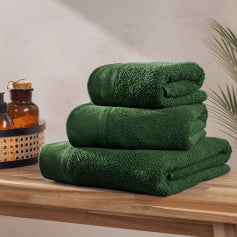 A selection of variously sized textured weave towels in a dark green shade, folded and arranged in a pile on a light wood surface.