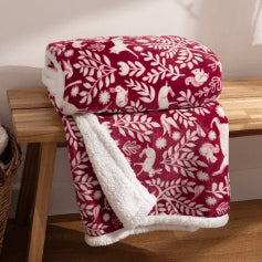 A berry red throw with a scandi-inspired printed design of white woodland animals and foliage, folded and presented on a light wooden bench.