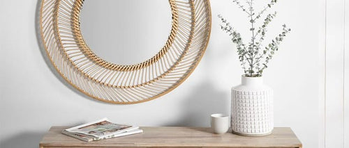 A round, bamboo woven mirror on a white wall, behind a wooden side table decorated with a book and houseplant.