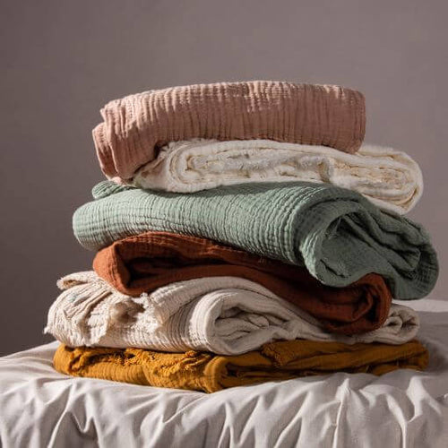 A selection of six crinkle cotton throws in various neutral and muted pastel shades, folded and piled on top of one another on a grey duvet cover.