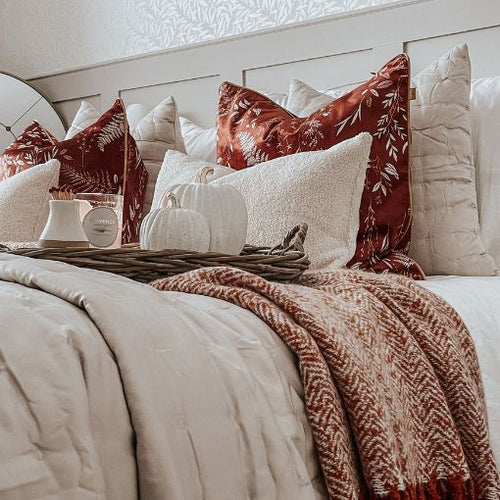 An autumn themed bed with rust red cushions, white cushions and a woven auburn throw, with a decorated serving tray.