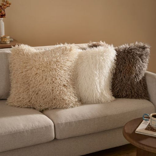 Three faux fur cushions on a sofa, in a beige, white and brown shade. All stacked up against a beige fabric sofa.
