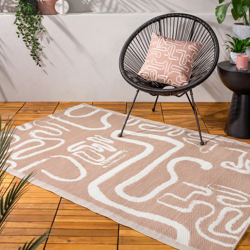 A neutral outdoor rug with a modern abstract design of swirling lines, laid on a wooden decking surface along with a black outdoor chair and a coordinating abstract outdoor cushion.